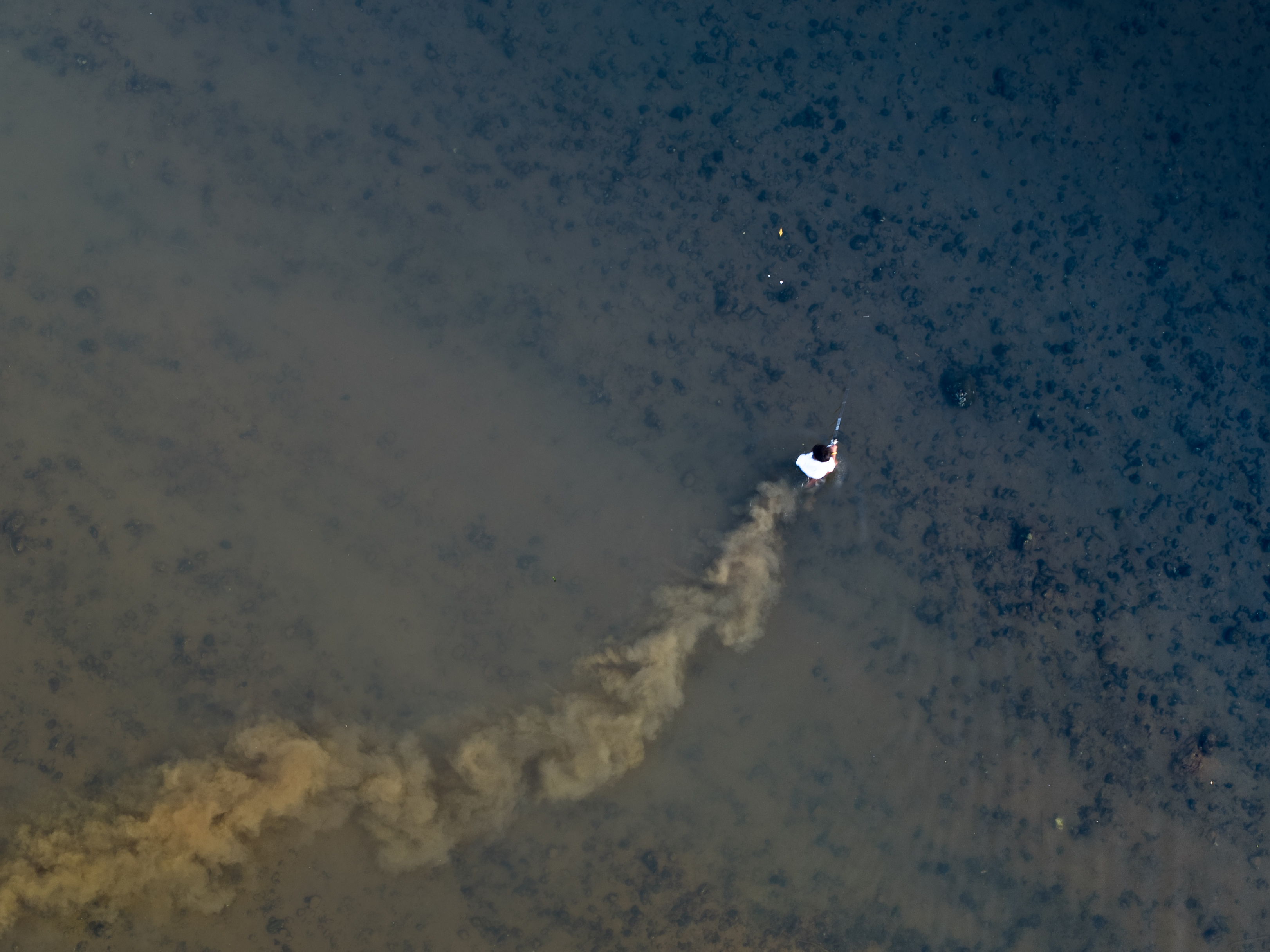 A person in a white and black jacket stands in shallow water with a trail behind them, possibly smoke.