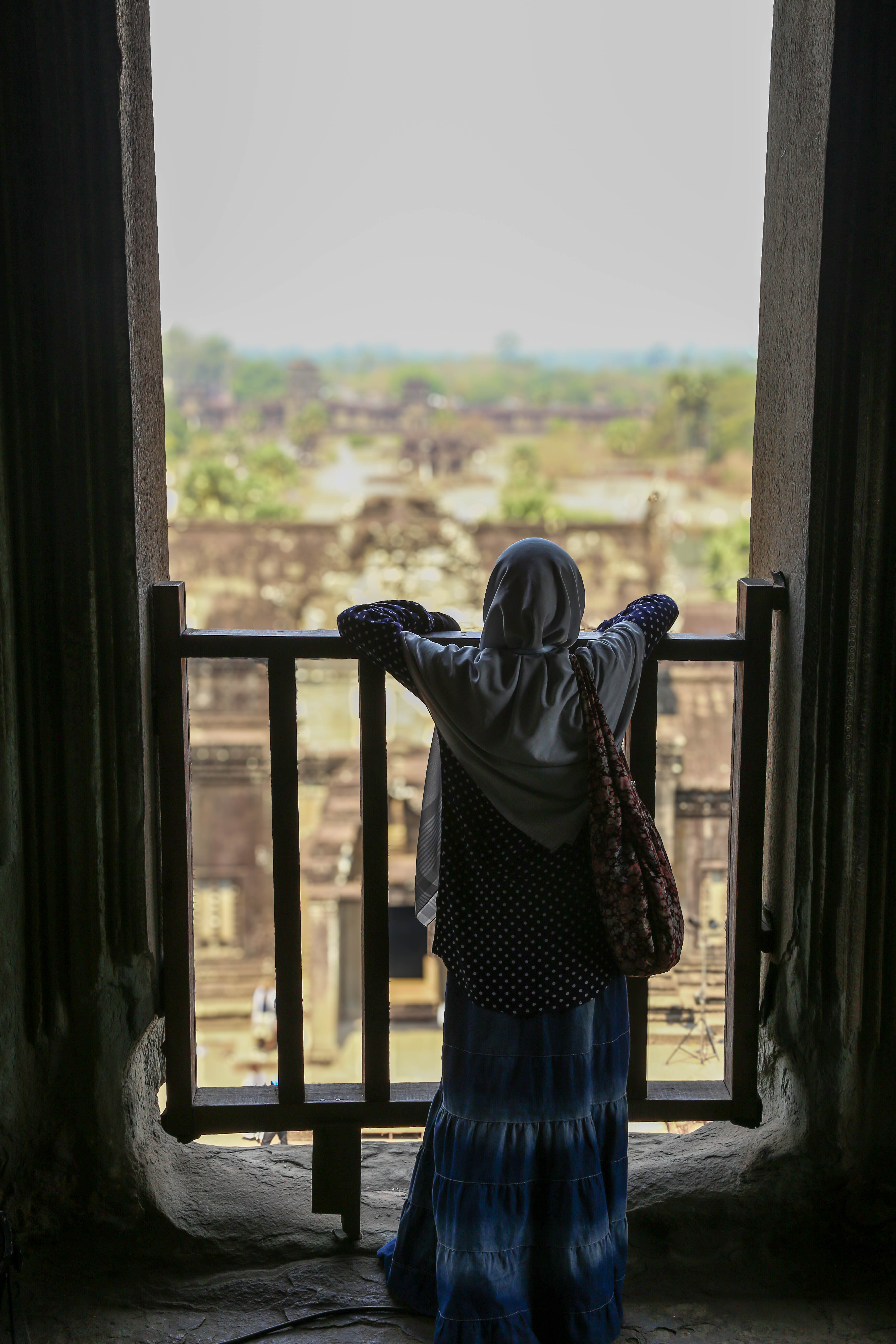 A female Muslim tourist, dressed in a long denim skirt, black and white polka dress, and grey headscarf, stands at a raili...